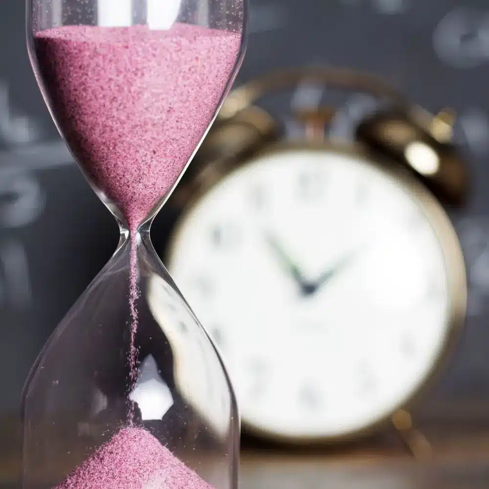 Close-up of an hourglass with pink sand falling, set against a blurred background of a vintage alarm clock, symbolizing time passing quickly.